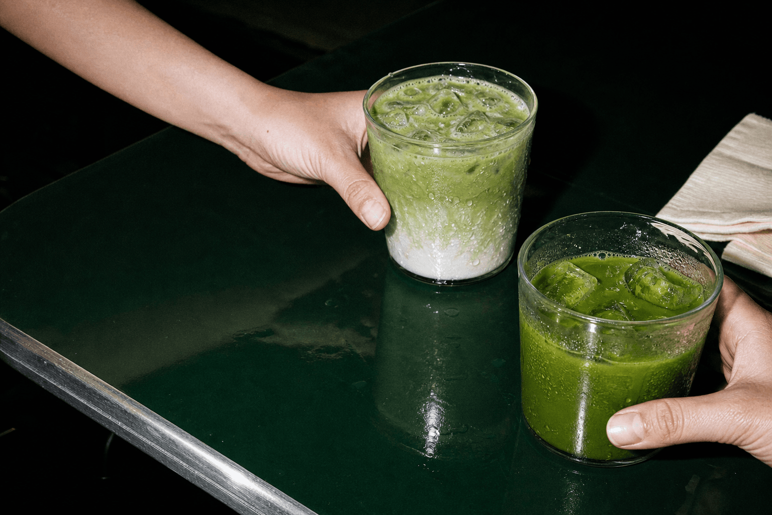 Two iced matcha drinks crossing a dark café table